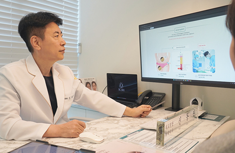 Asian doctor in a white coat explains information to a patient at a clinic desk, pointing to a computer monitor on the desk.