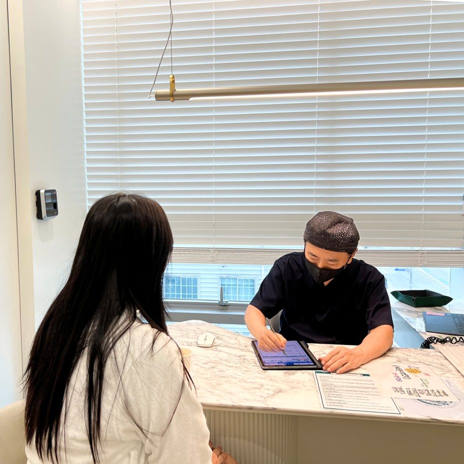 A doctor in navy scrubs and a mask sits at a marble reception desk, using a tablet to review information while a patient sits opposite.